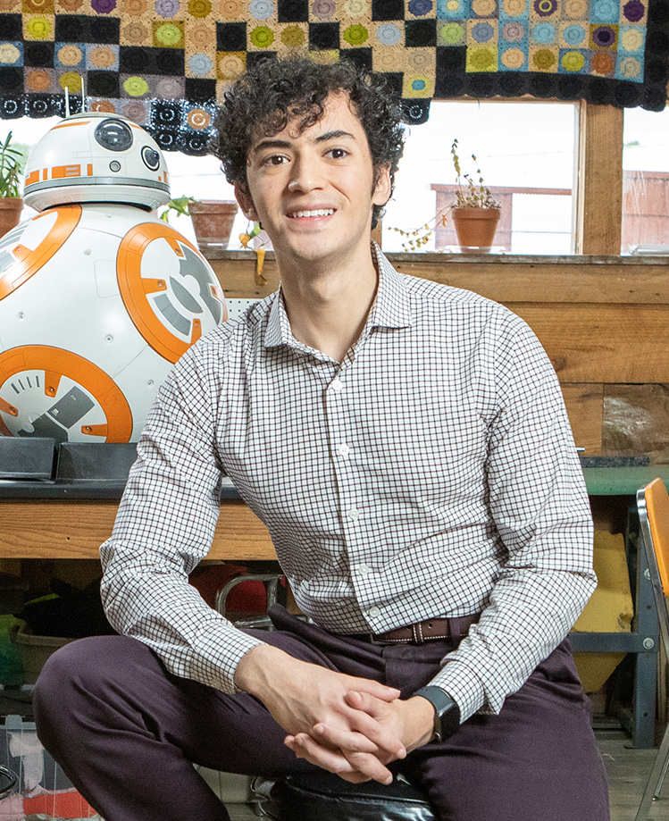 A photo of Matias Silva sitting in a lab with a droid in the background.